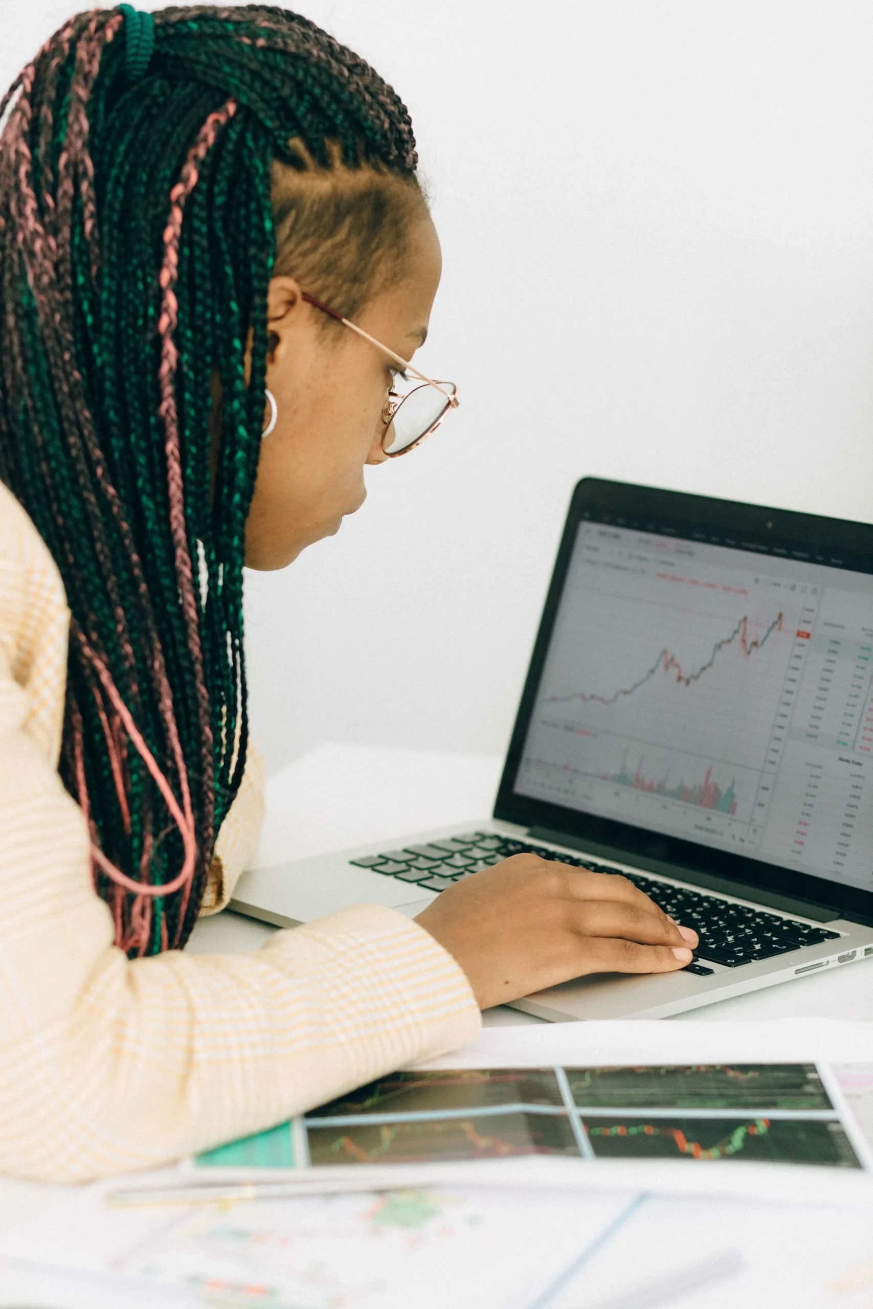 A person is working on a laptop, analyzing financial charts and data displayed on the screen. The individual is likely engaging in trading or financial planning, surrounded by a calculator and various documents on the desk, reflecting a focused and analytical environment.